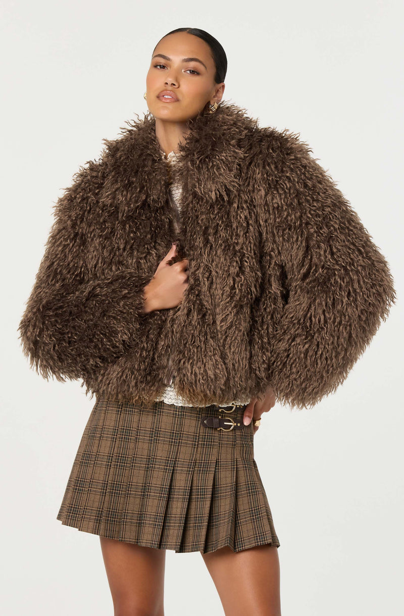 A woman is wearing a large, fluffy, dark brown faux fur coat that covers her shoulders and upper body. The coat has a high collar and appears soft and plush. She is also wearing a brown plaid pleated skirt with a black belt featuring silver details.