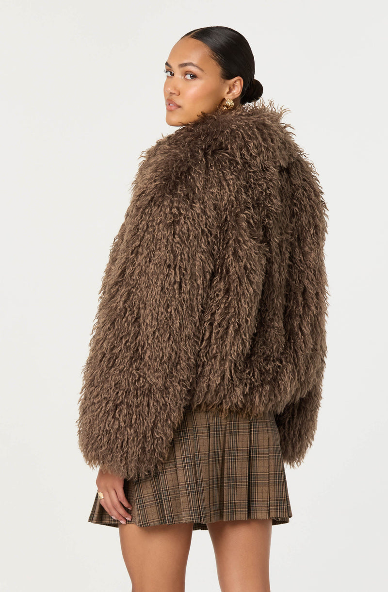 A woman is wearing a large, fluffy, brown faux fur coat with a textured, curly appearance. She is also wearing a pleated skirt with a classic plaid pattern in shades of brown and black.