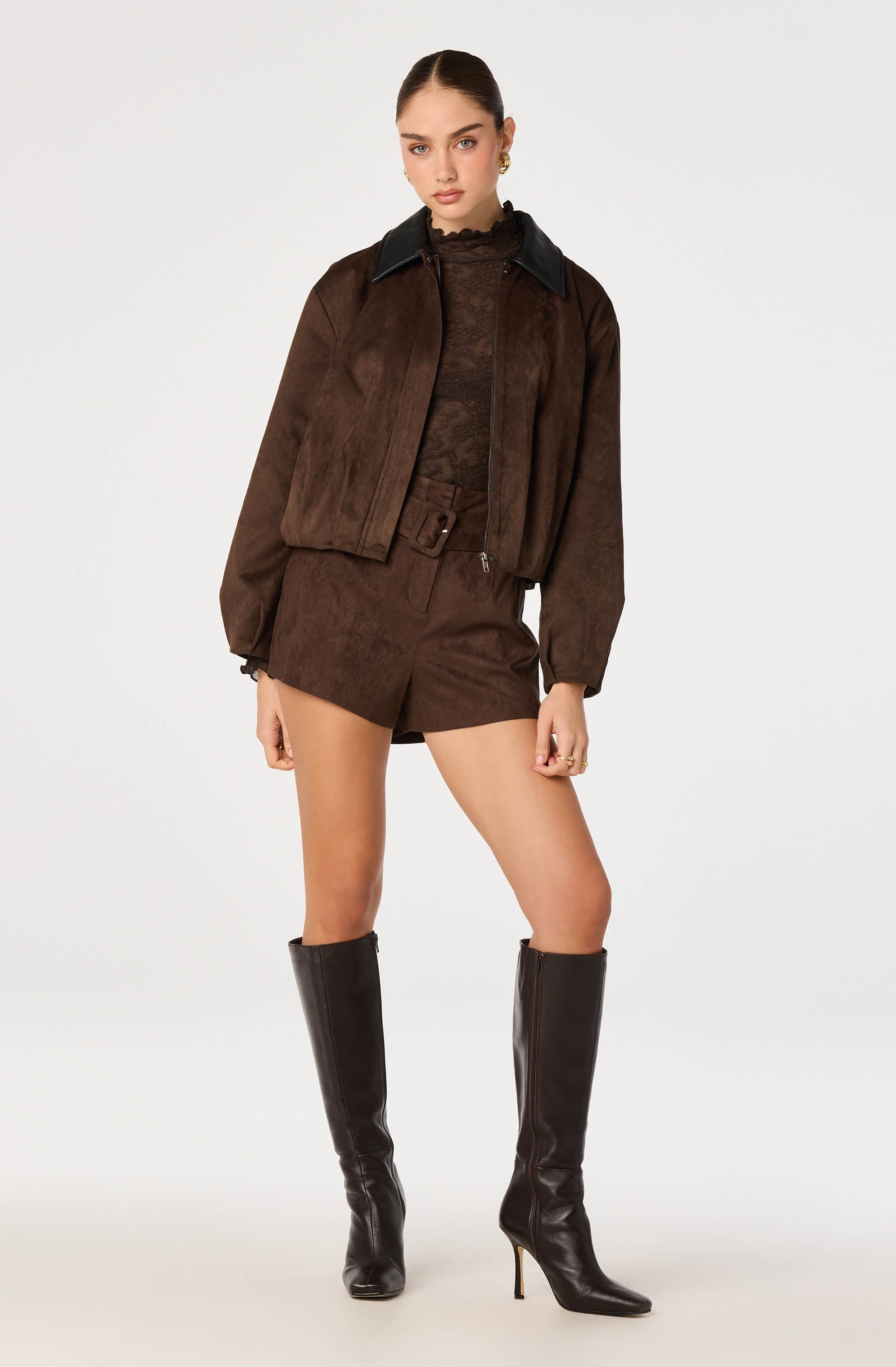 Young woman wearing a brown suede bomber jacket with black collar, matching shorts, and black knee-high high-heeled boots.