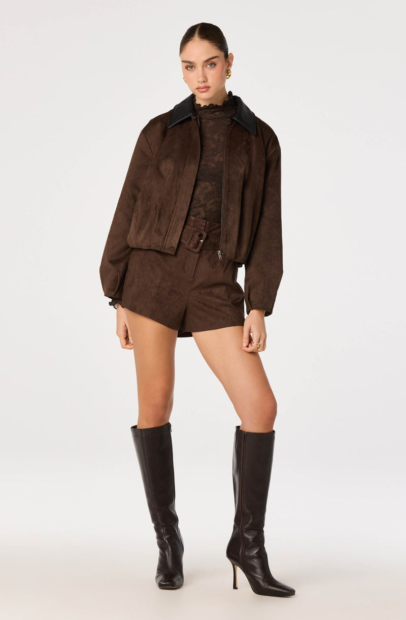 Young woman wearing a brown suede bomber jacket with black collar, matching shorts, and black knee-high high-heeled boots.
