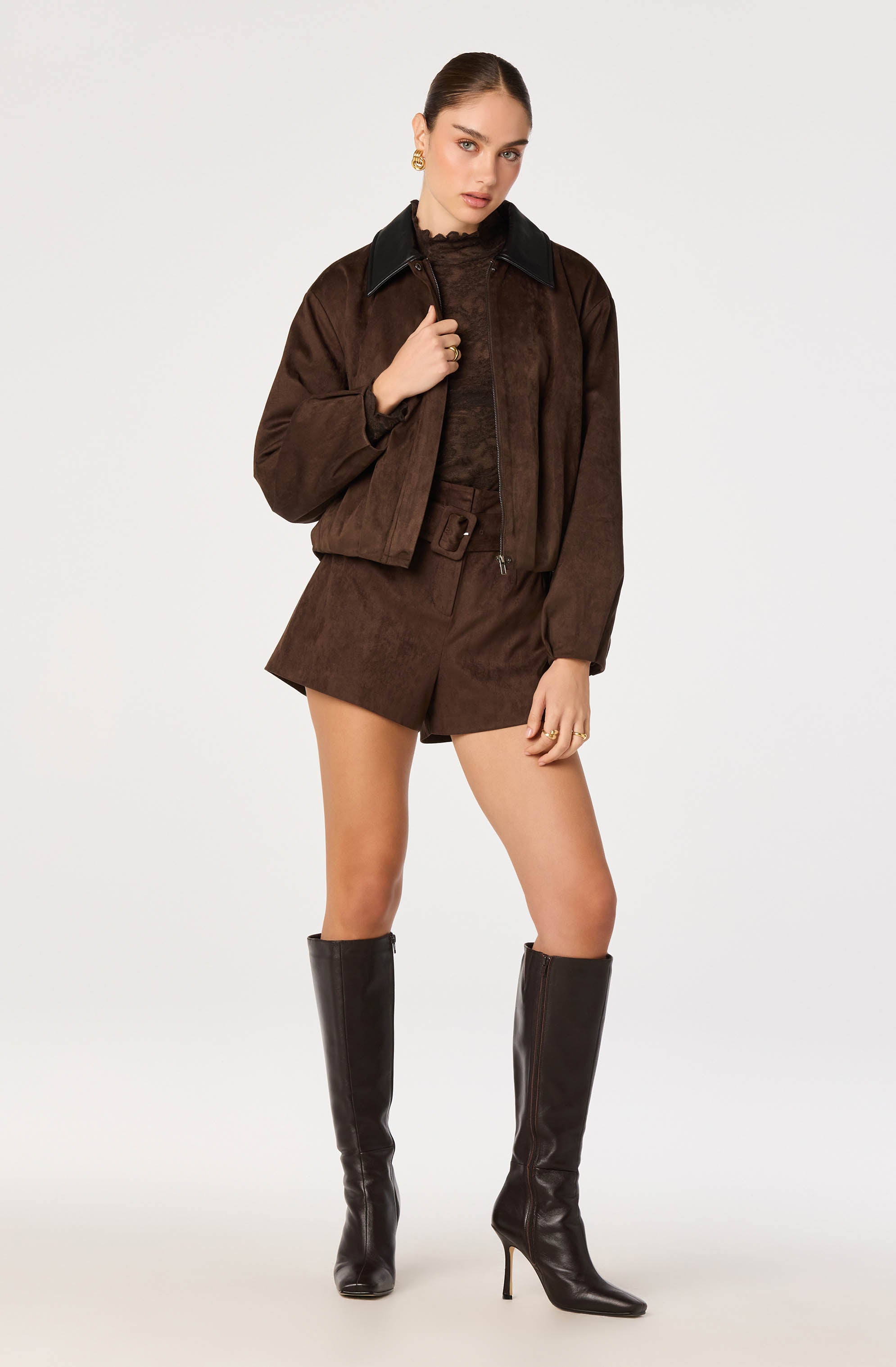 Woman wearing a brown suede jacket with a black collar, matching brown shorts, and tall black high-heeled boots.