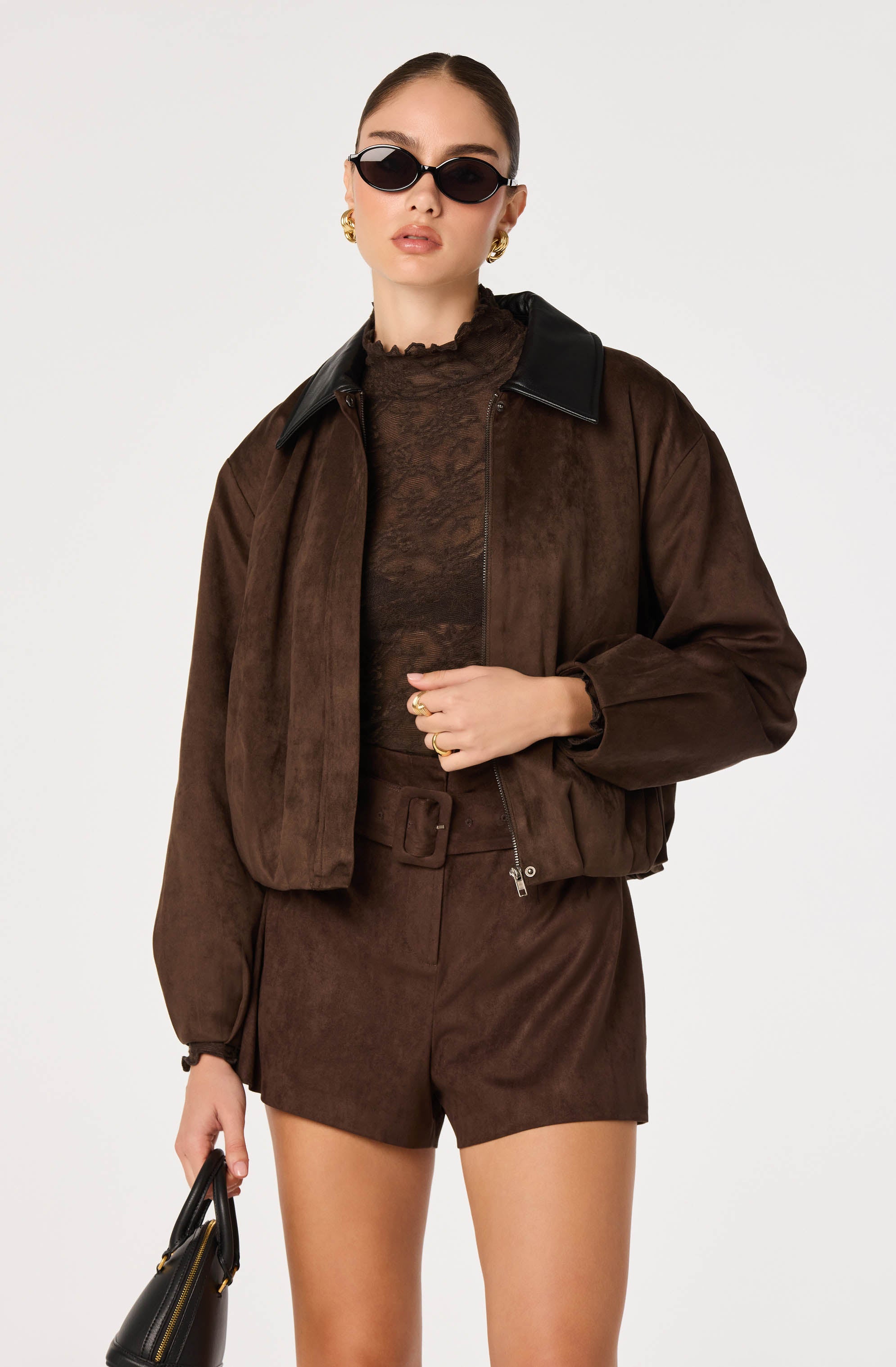 Woman wearing a brown suede jacket, matching high-waisted shorts, black lace top, black sunglasses, and gold jewelry.