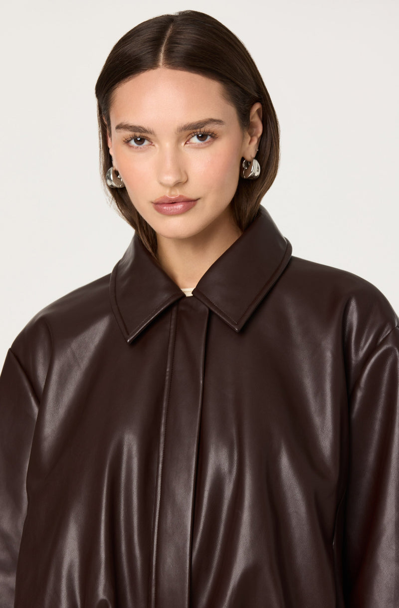 A young woman with shoulder-length brown hair is wearing a dark brown, shiny leather jacket with a pointed collar. She has on large, silver hoop earrings and a subtle makeup look. The background is plain and light-colored.