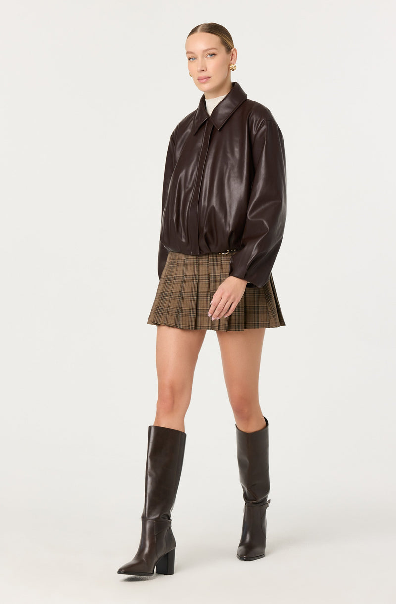 A woman is wearing a dark brown, shiny leather bomber jacket with a wide collar and elastic cuffs. She has paired it with a plaid mini skirt in shades of brown and beige, and tall dark brown leather boots with high block heels.