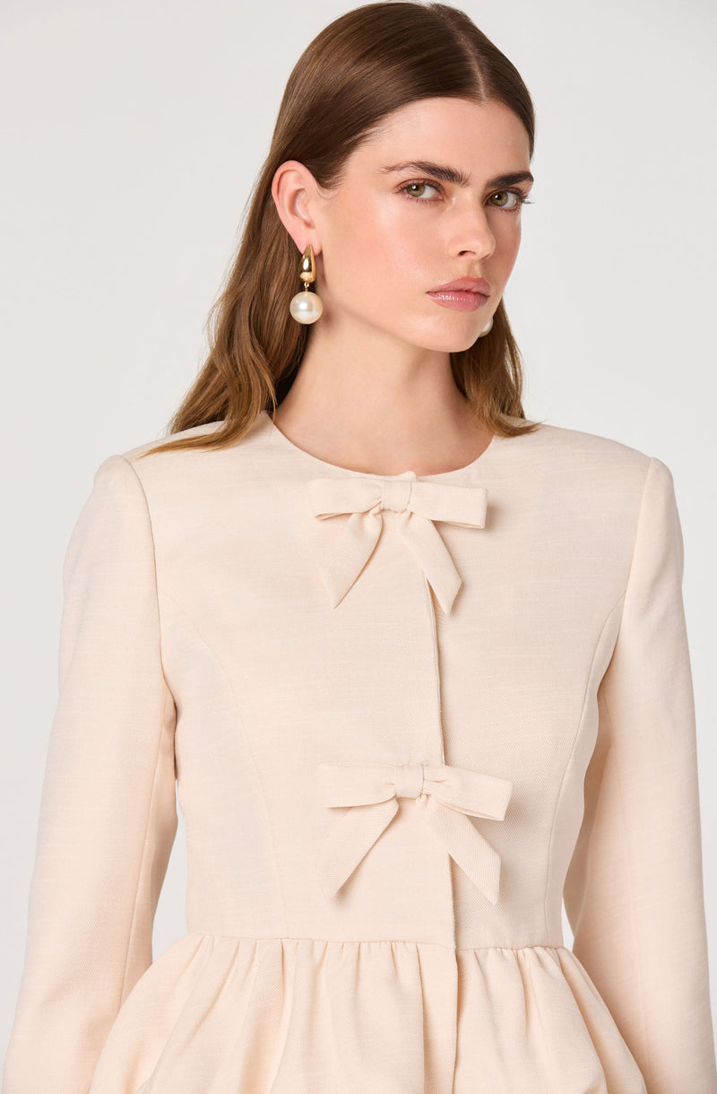 Woman wearing a cream-colored dress with two bow details on the front and a gathered waist.