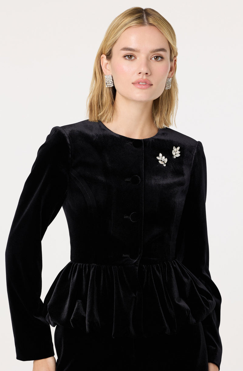 A woman is wearing a black velvet jacket with a round neckline and long sleeves. The jacket has decorative leaf-shaped embellishments on the left side of the chest and features a ruched, gathered waistline that creates a peplum effect.