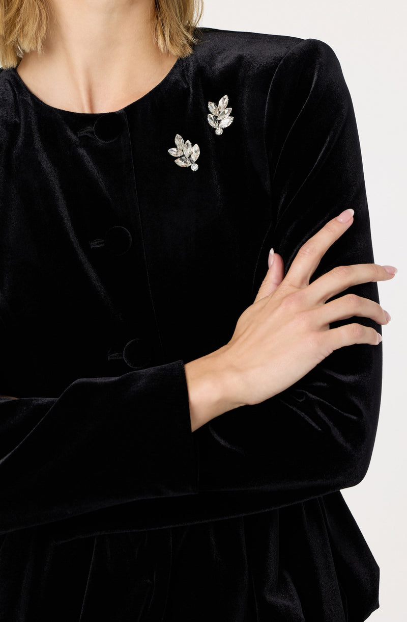 A woman is wearing a black velvet jacket with a round neckline. The jacket has a row of large, round buttons down the front. On the left side of the jacket, near the shoulder, there are two decorative brooches in the shape of leaves, adorned with shiny, clear stones. The woman's left hand is crossed over her body, resting on her right arm. The fabric of the jacket has a soft, plush texture, and the overall style appears elegant and sophisticated.