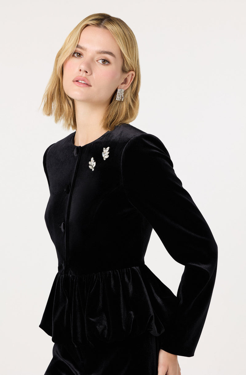 A woman is wearing a black velvet jacket with a round neckline and long sleeves. The jacket features decorative embellishments near the collar, including two brooches with a floral or leaf design. The waist of the jacket is gathered, creating a ruffled effect at the hem. She has accessorized with large, square, rhinestone earrings.