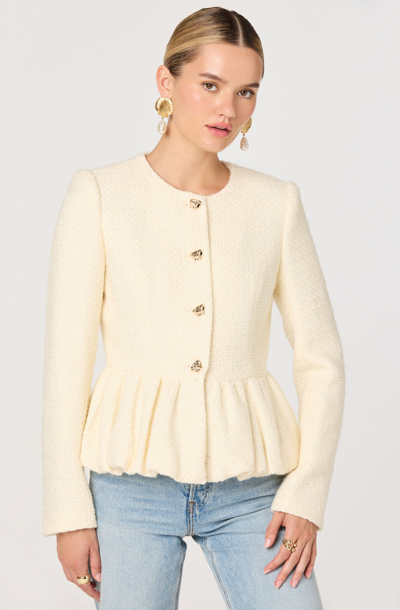 A woman is wearing a cream-colored textured jacket with a round neckline and long sleeves. The jacket features decorative buttons down the front and a gathered peplum hem. She pairs it with light-wash jeans.