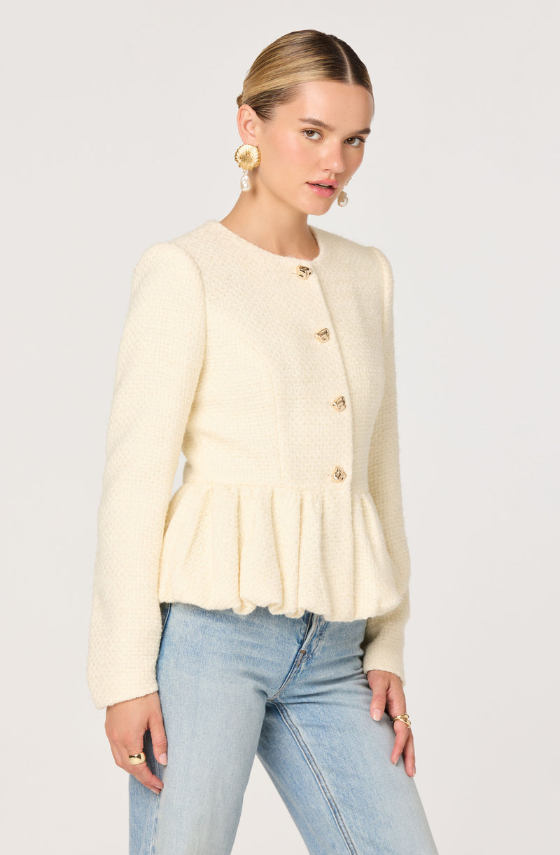 A woman is wearing a cream-colored, textured, long-sleeve jacket with gold buttons down the front. The jacket features a peplum-style ruffle at the waist, creating a gathered effect. She has paired it with light-wash denim jeans. She is also wearing large, gold, circular earrings and gold rings on her fingers.