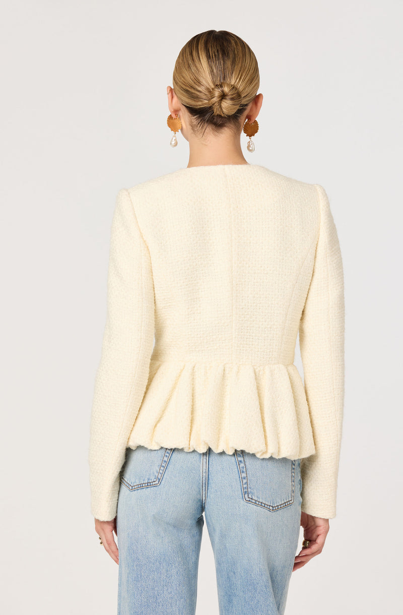 A woman is wearing a cream-colored textured blazer with a peplum hem and slightly padded shoulders. She has on light blue high-waisted jeans with visible back pockets. Her hair is styled in a low bun, and she is accessorized with gold hoop earrings that have dangling pearl accents.