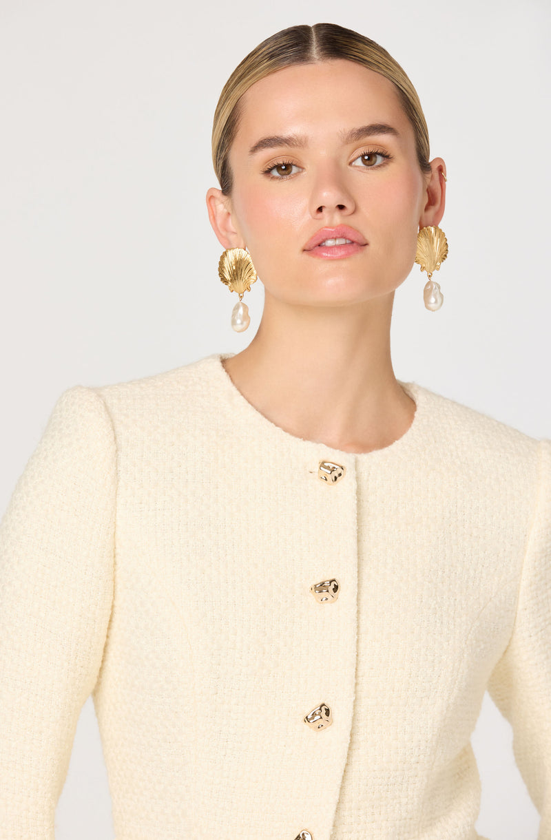 A woman is wearing a cream-colored, textured, long-sleeved jacket with a round neckline. The jacket features a row of gold-tone buttons down the front with a decorative design. She accessorizes with large gold statement earrings that have a shell shape and dangling pearl drops. Her hair is styled sleek and pulled back.