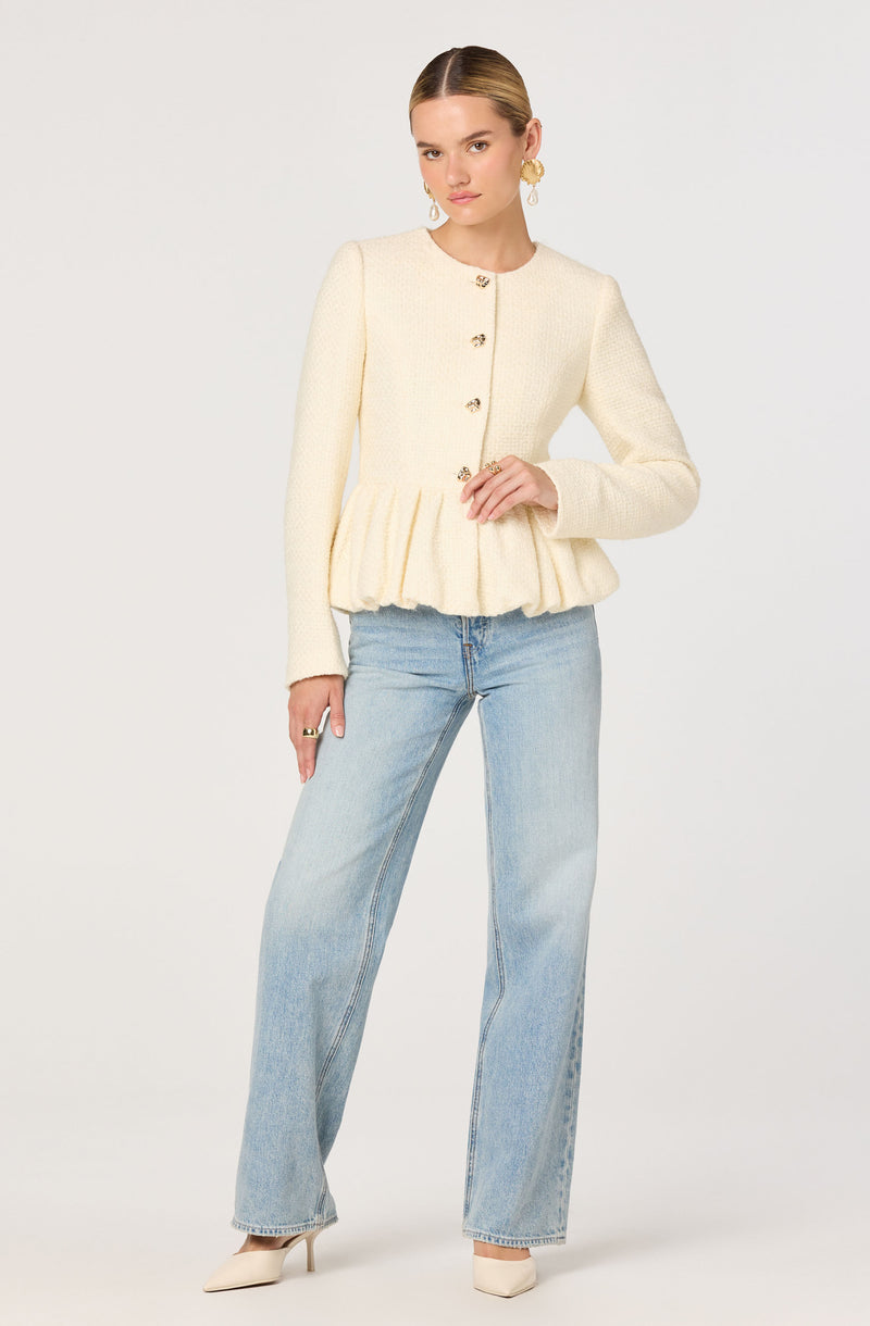 A woman is wearing a cream-colored, textured knit jacket with a round neckline and long sleeves. The jacket features a row of decorative buttons down the front and has a gathered, ruffled hem that creates a peplum effect. She pairs it with light-wash, high-waisted, wide-leg jeans and white pointed-toe heels. She is accessorized with gold hoop earrings and rings.
