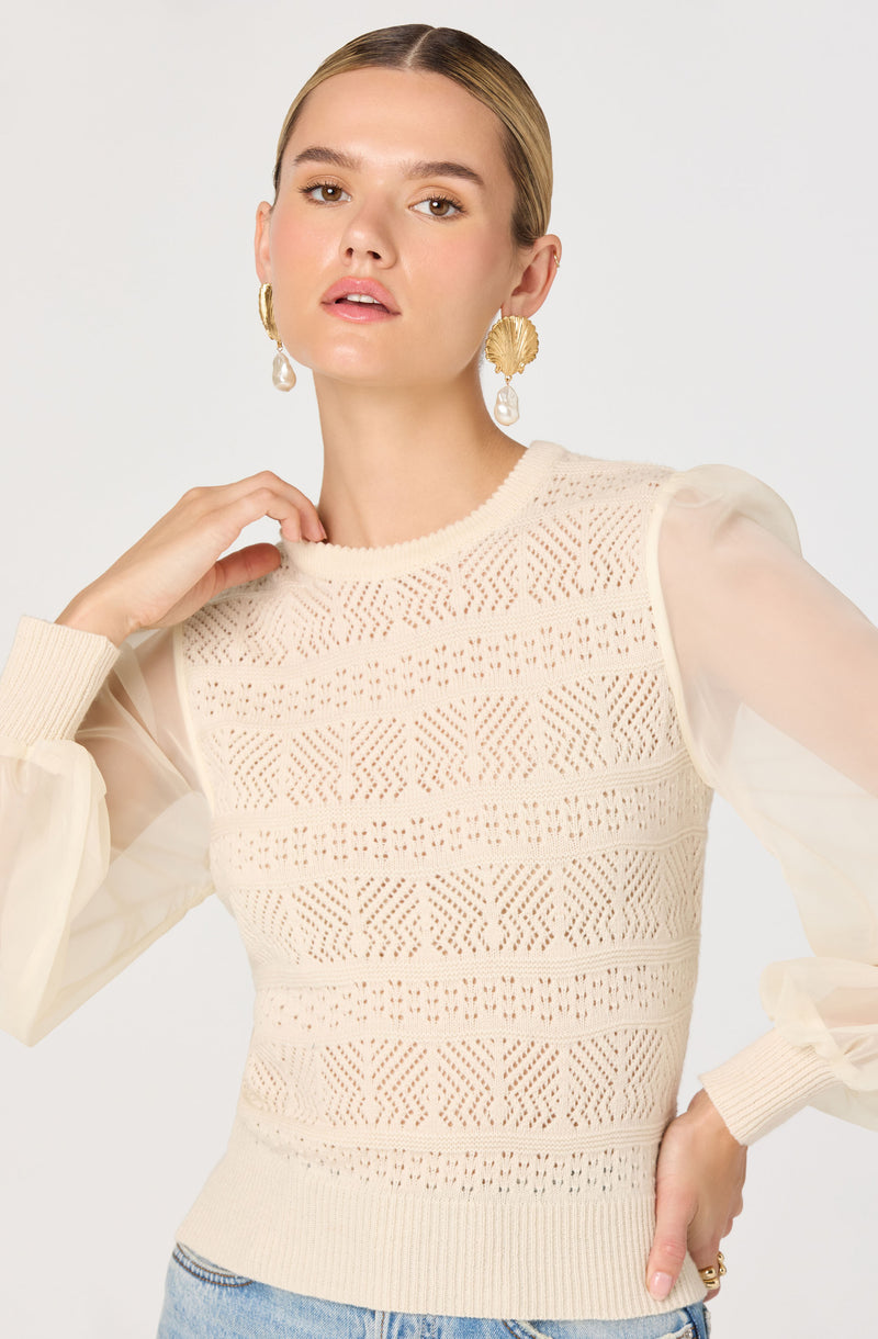 A woman is wearing a cream-colored sleeveless knit top with intricate geometric lace patterns throughout. The top features a round neckline and appears to be layered over sheer, puffed long sleeves. She is accessorizing with large, gold statement earrings that have a fan shape and dangling pearl details.