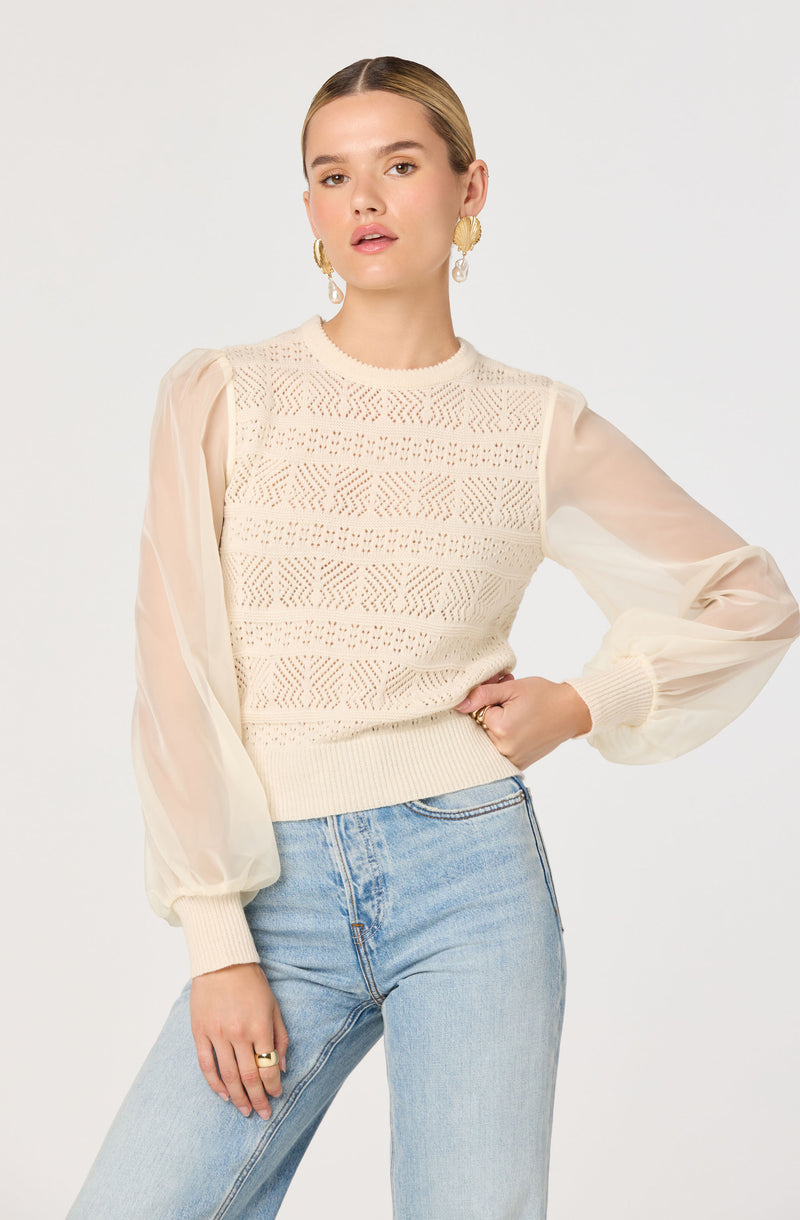 The woman is wearing a cream-colored sweater with intricate knitted patterns on the front. The sweater features sheer, long, puffed sleeves made of a light, translucent fabric, gathered at the cuffs with ribbed knit. The body of the sweater is fitted at the waist with a ribbed hem. She pairs it with light blue high-waisted jeans.