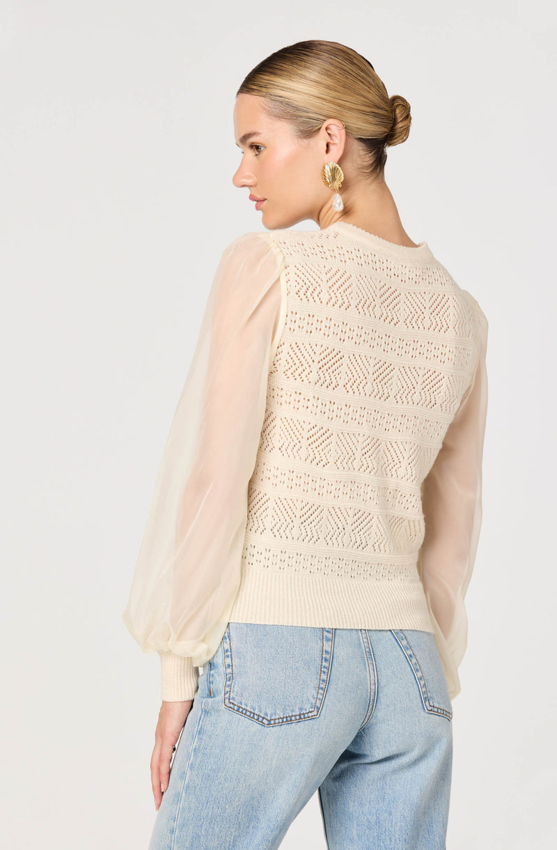 A woman is wearing a cream-colored knit sweater with intricate openwork patterns on the back, paired with sheer, pale beige long-sleeve chiffon sleeves. She has light blue jeans and is accessorized with large gold earrings featuring white pendants. Her hair is styled in a neat low bun.