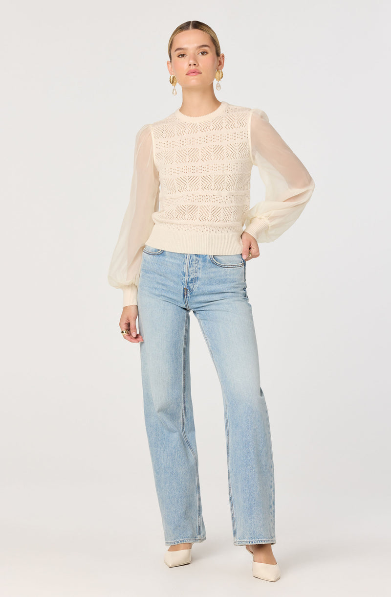 A woman is wearing a cream-colored, textured knit sweater with sheer, puffed long sleeves. She pairs it with light-wash, high-waisted, straight-leg jeans and beige pointed-toe shoes. She has large, gold hoop earrings and is standing against a plain white background.