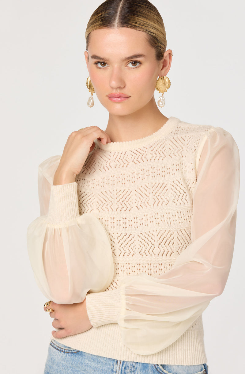 A woman is wearing a cream-colored sweater with intricate knit patterns on the body. The sweater has sheer, puffed long sleeves made of a lightweight, translucent fabric. She is also wearing large gold earrings with pearl accents.