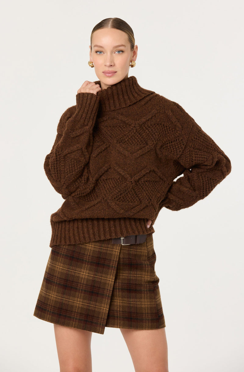 A woman is wearing a chunky, oversized brown knit sweater with a high turtleneck collar, featuring a textured cable pattern. She has paired it with a brown plaid skirt with red and yellow accents, and a black belt. She is also wearing gold earrings.