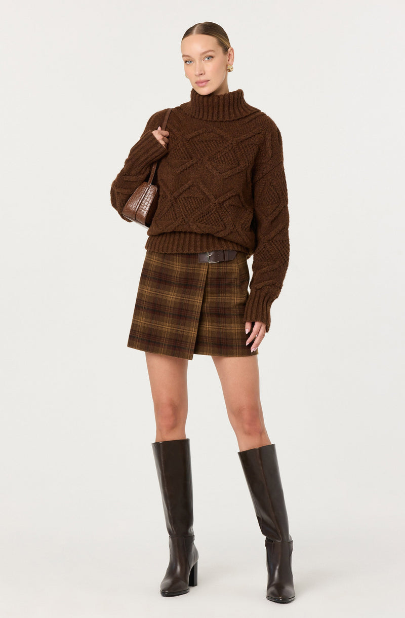 A woman is wearing a chunky, brown, cable-knit turtleneck sweater with long sleeves. She has a plaid A-line skirt in shades of brown, green, and yellow, paired with knee-high black leather boots with a block heel. She is also carrying a small brown bag over her shoulder.