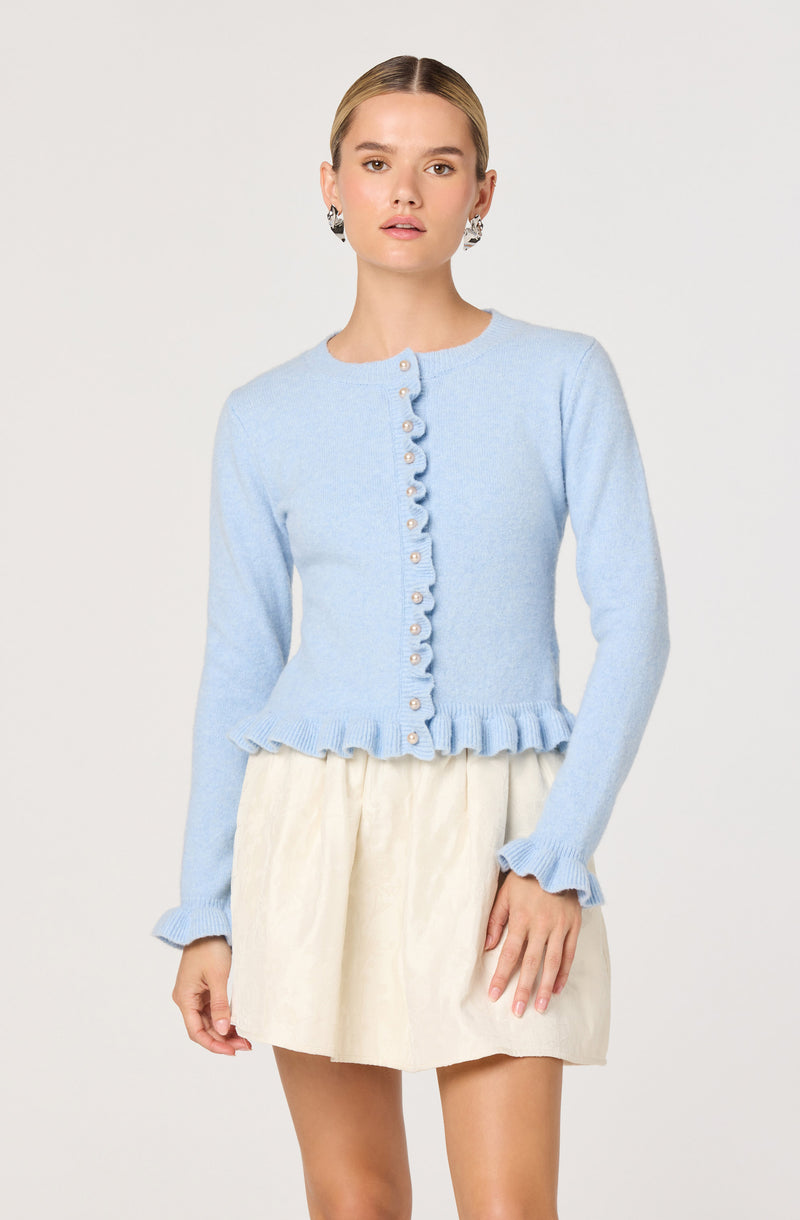 A woman is wearing a light blue, long-sleeved knit cardigan with pearl buttons down the front. The cardigan features a ruffled trim along the hem and cuffs. She pairs it with a knee-length, off-white skirt. She has her hair pulled back and is accessorized with large, black and white earrings.