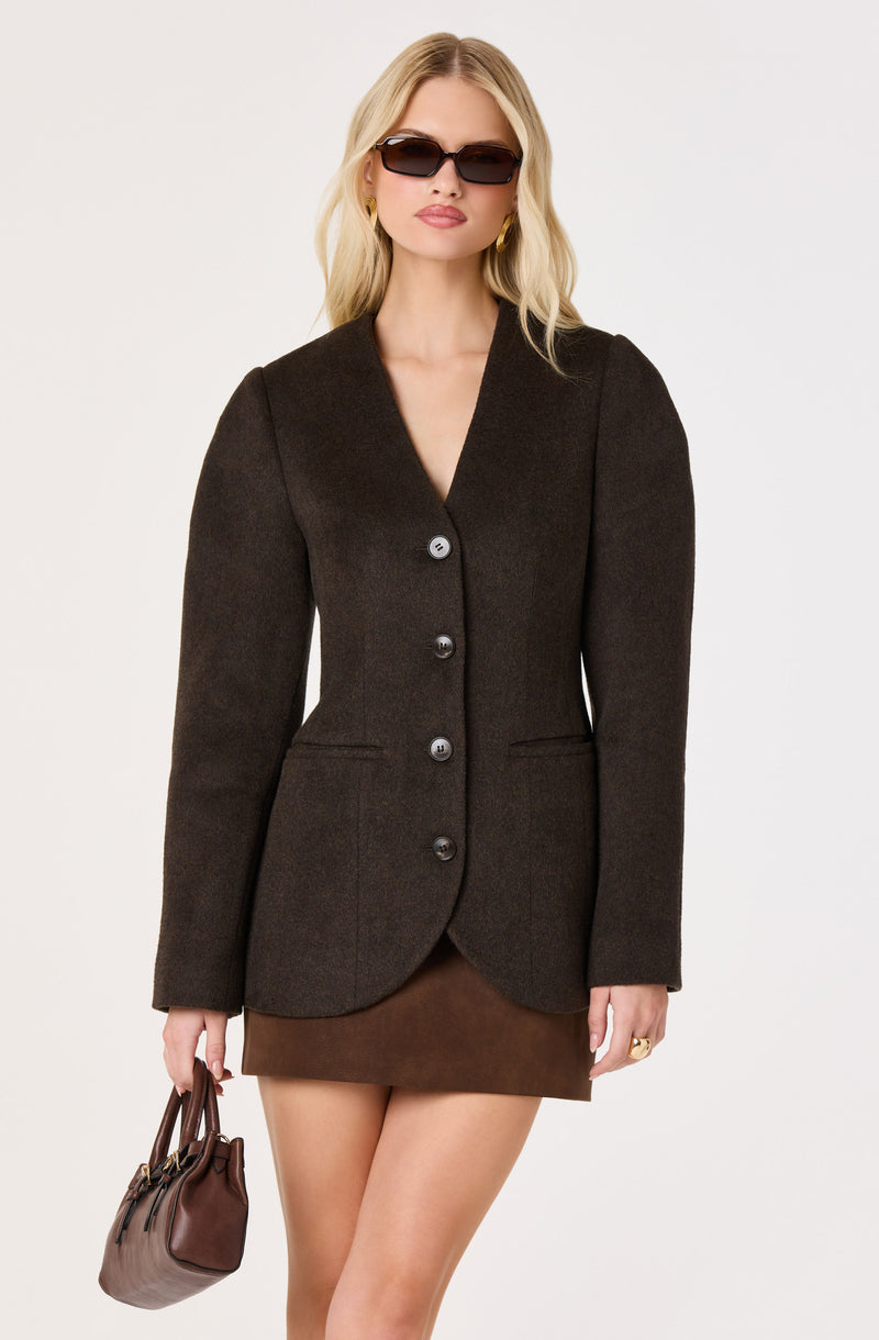Woman wearing a dark blazer with large buttons, brown skirt, black sunglasses, and gold earrings.