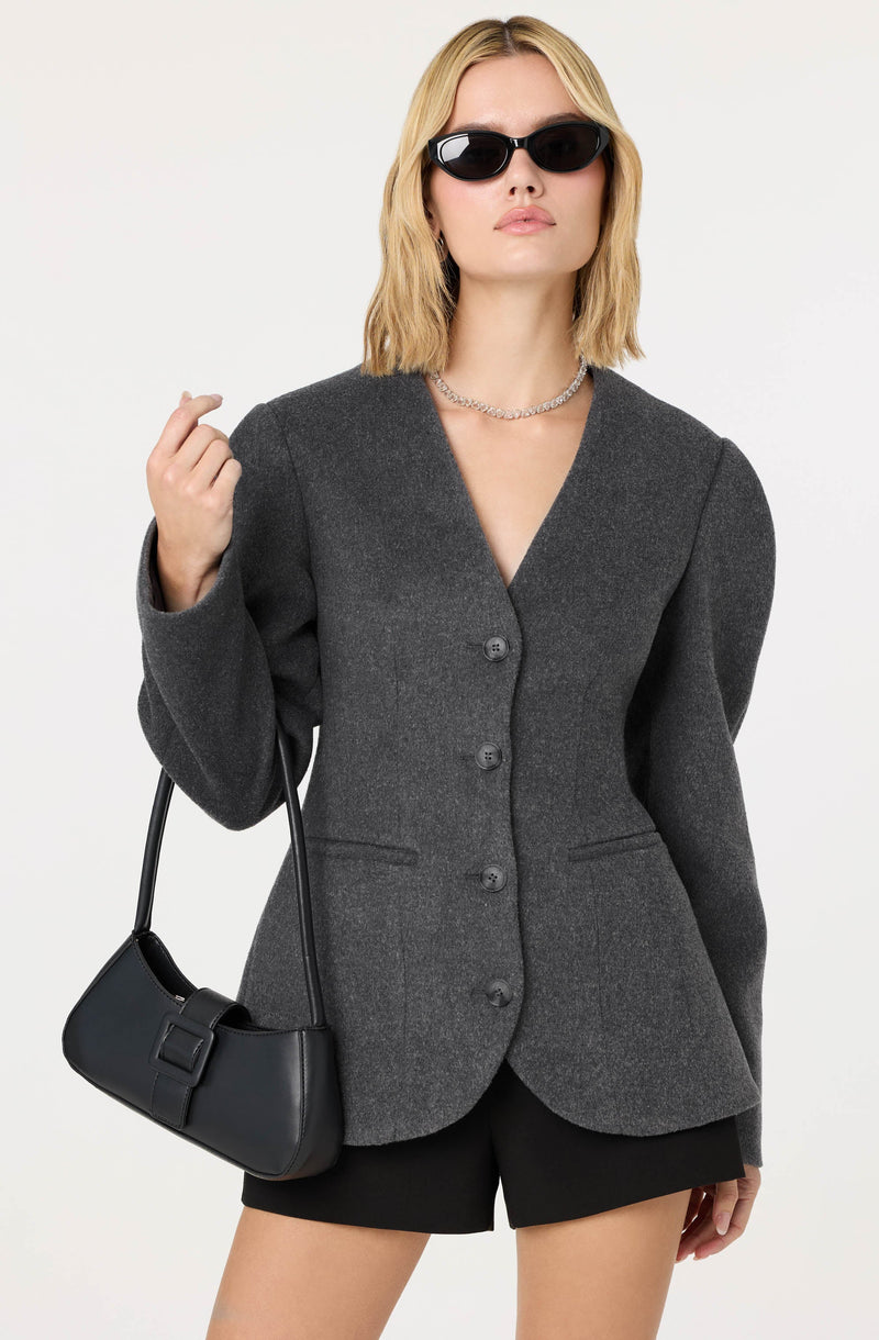 A woman wearing a fitted, dark gray blazer with a v-neckline and four buttons down the front. The blazer has a slightly textured fabric and features small pockets on each side. She is also wearing black shorts, sunglasses, a silver necklace, and is carrying a small black handbag with a buckle detail.