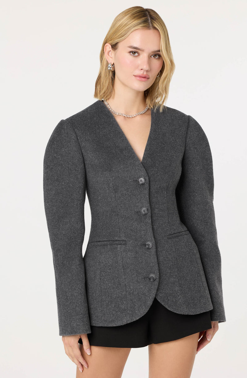 A woman is wearing a tailored, dark gray blazer with a fitted silhouette, structured shoulders, and long sleeves. The blazer has a notched lapel collar and four buttons down the front. She pairs it with black shorts and accessorizes with a silver necklace and matching earrings.