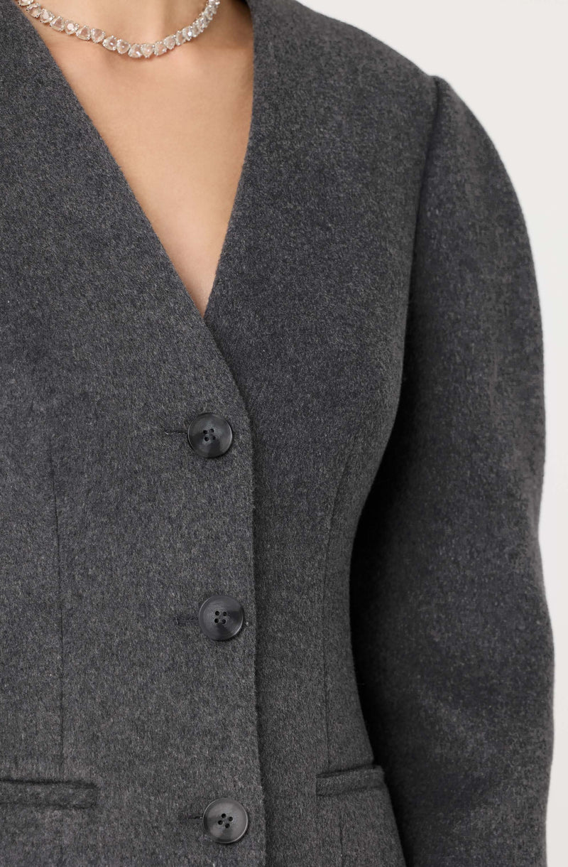 A person is wearing a dark gray tailored blazer with a V-shaped neckline. The blazer features three visible black buttons down the front and small, discreet slit pockets on each side. The individual accessorized with a pearl necklace.