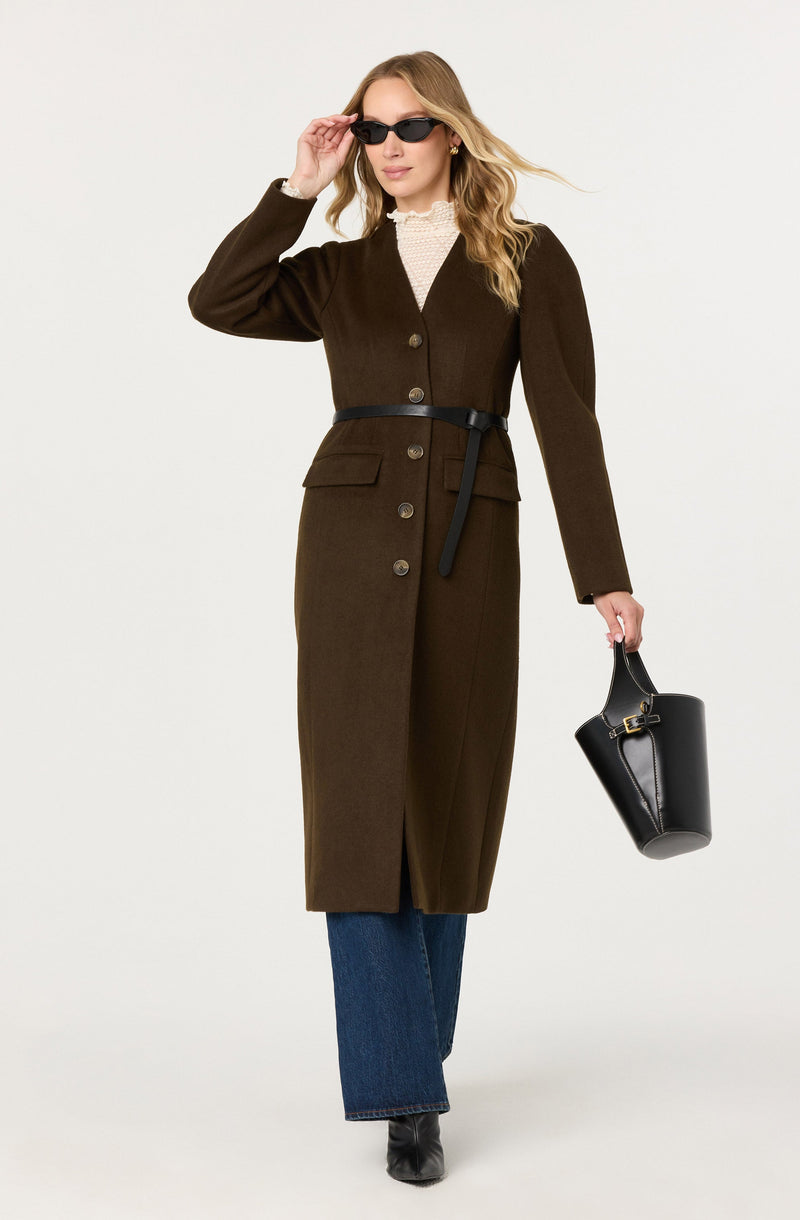 The woman is wearing a long, dark brown coat with a V-neckline, button closure down the front, and a matching belt tied around her waist. Underneath, she has a cream-colored, textured top with a high neckline. She is also wearing dark sunglasses, large earrings, blue wide-leg jeans, and black boots. She carries a black bucket bag with a gold buckle and has long, wavy blonde hair.