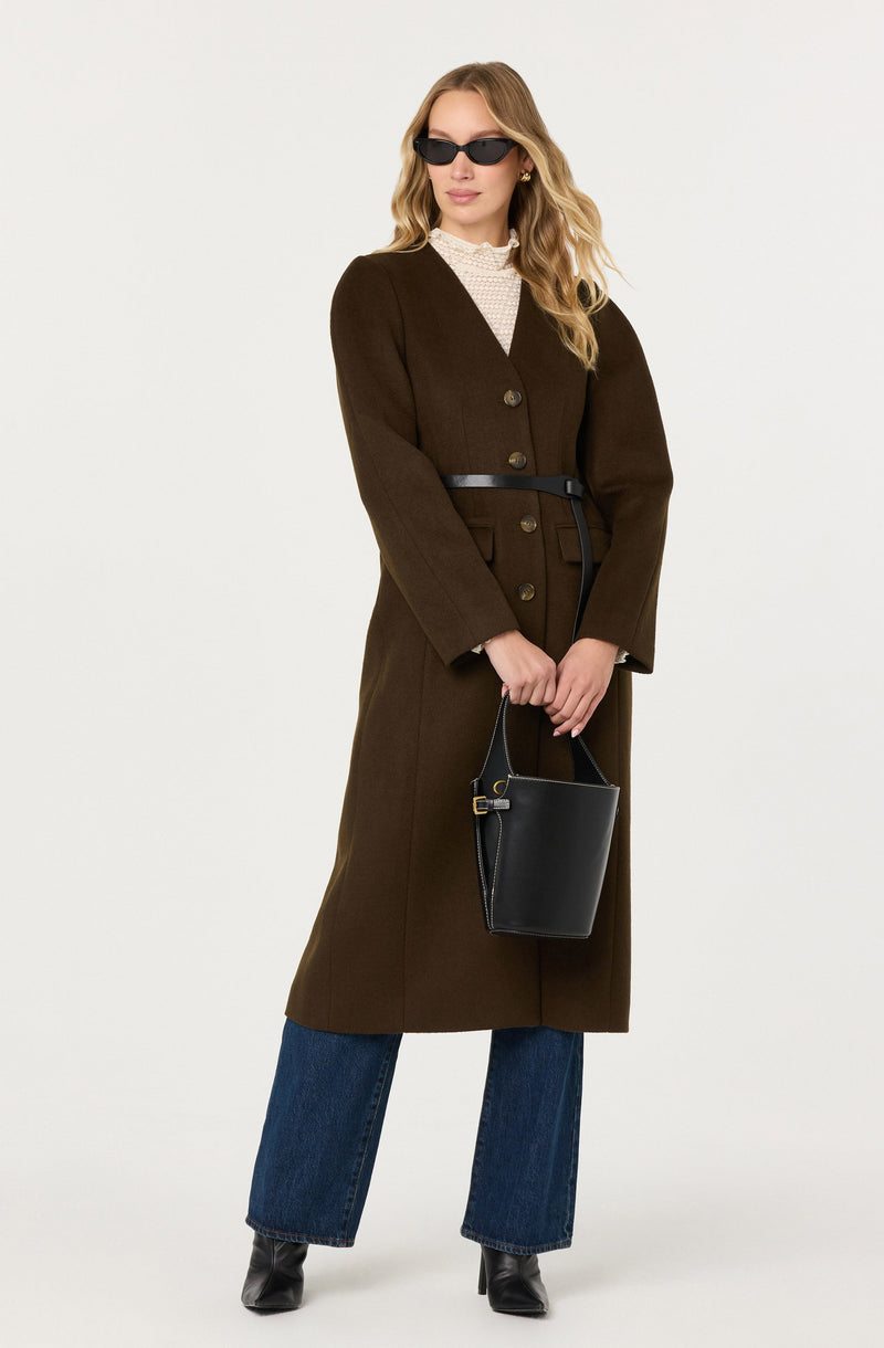 A woman is wearing a long, dark brown coat with large buttons down the front and a belted waist. Underneath, she has a cream-colored, high-neck blouse with a textured pattern. She is accessorized with black sunglasses and small earrings, and she is holding a black handbag with a strap. She is also wearing dark blue wide-leg jeans and black heeled boots.