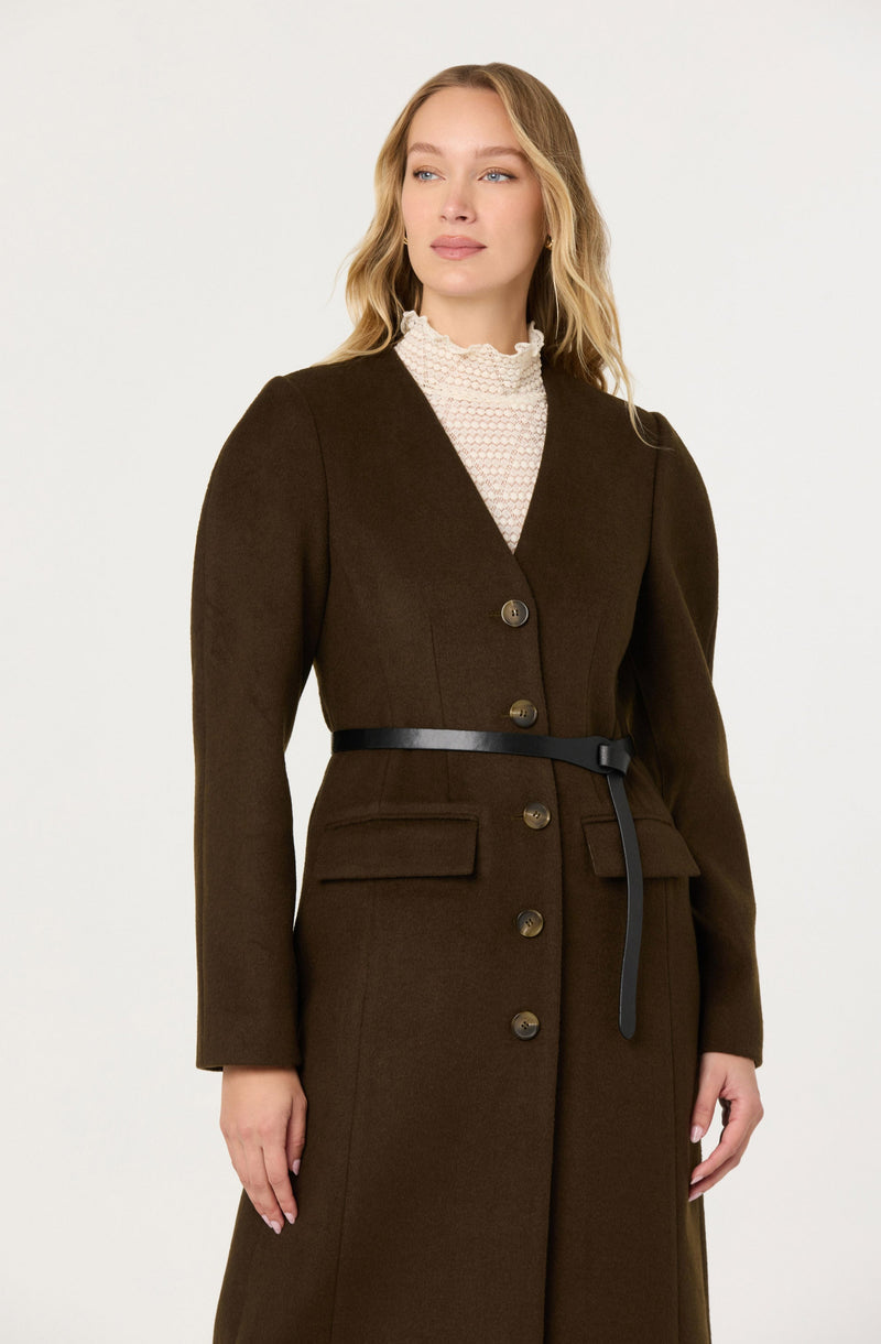 A woman is wearing a long, dark brown coat with a tailored fit and a row of buttons down the front. The coat has two flap pockets on the front and is accessorized with a thin black belt tied at the waist. She has on a high-neck, cream-colored, textured blouse with a ruffled collar underneath the coat.