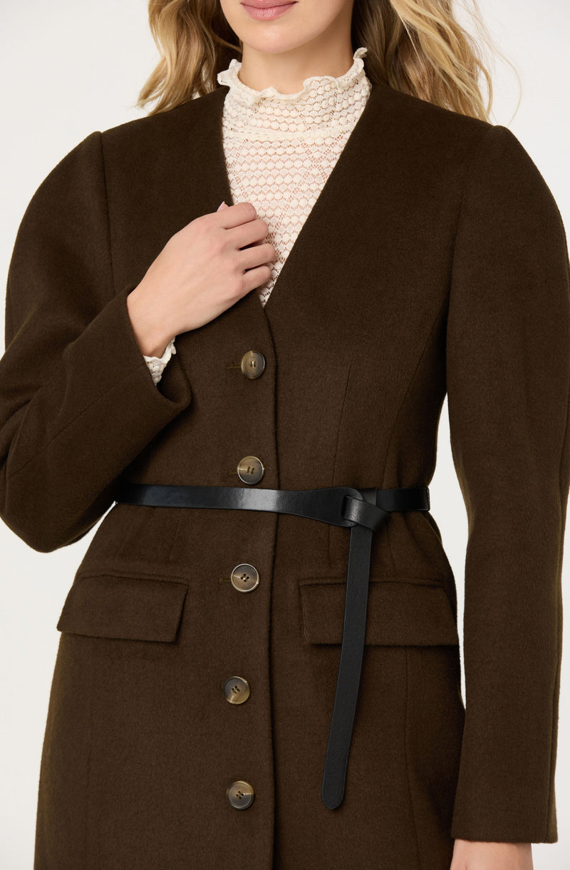 A woman is wearing a dark brown, tailored coat with large buttons down the front and flap pockets on each side. The coat has a black belt tied around the waist. She also has on a white, textured, high-neck blouse with a ruffled collar underneath the coat.