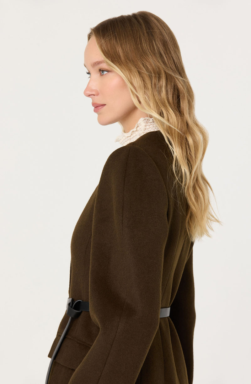 A woman is wearing a dark brown, tailored coat with a belt at the waist. The coat has a smooth, structured fit and long sleeves. She is also wearing a high-neck, lace-trimmed top underneath the coat.