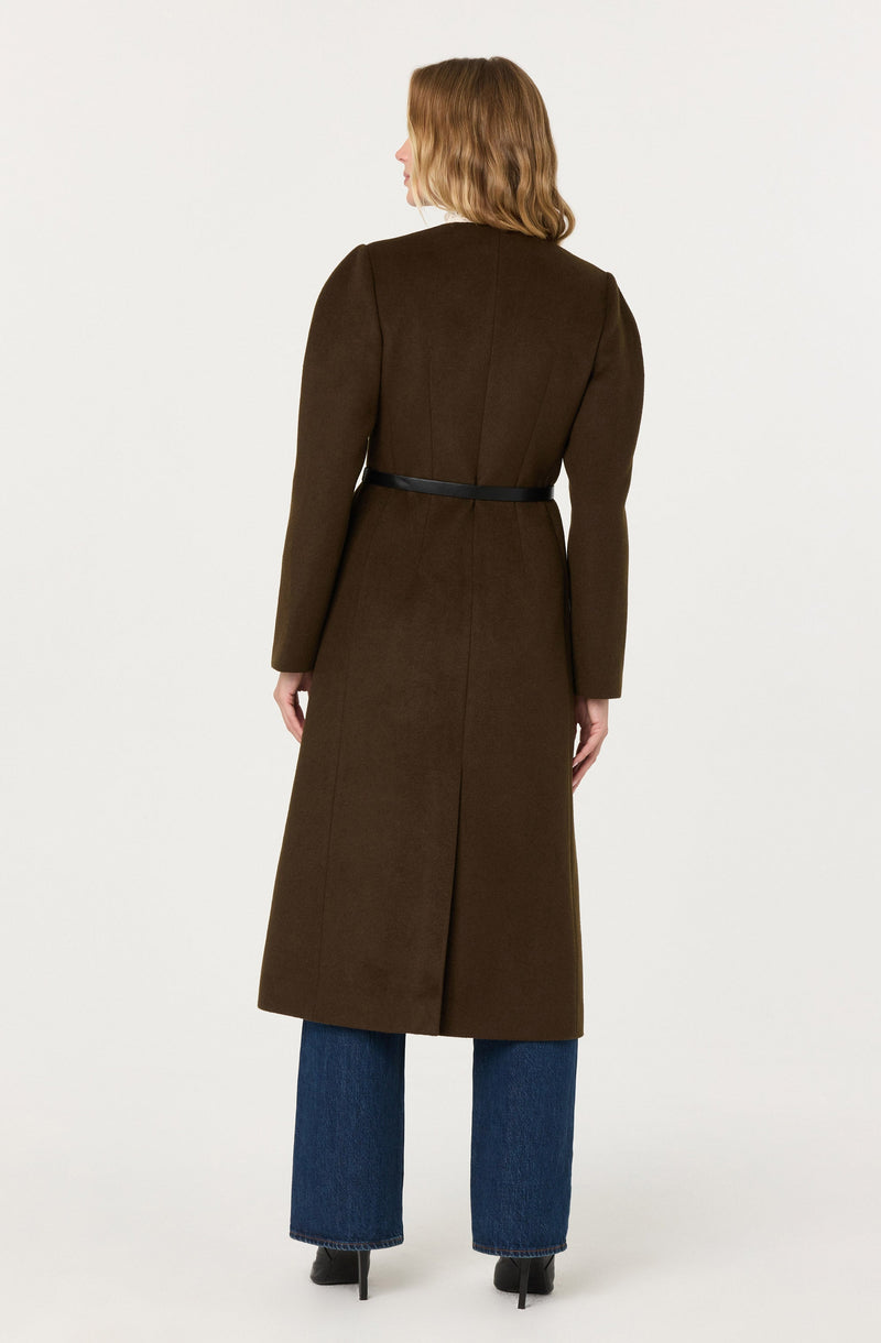 The image shows a woman wearing a long, brown coat that reaches below her knees. The coat features a black belt around the waist, long sleeves, and a tailored fit. She is also wearing wide-leg blue jeans and black high-heeled shoes. Her hair is shoulder-length and light brown.