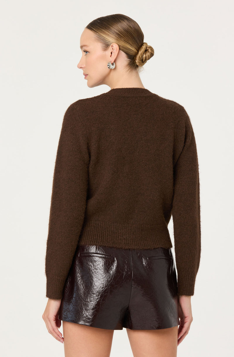 A woman is wearing a cozy, dark brown, long-sleeved sweater with a round neckline. The sweater appears soft and has a relaxed fit with ribbed cuffs and hem. She pairs it with high-waisted, shiny black leather shorts that have a smooth texture and a tailored fit.