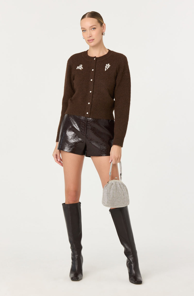 A woman is wearing a dark brown, long-sleeved, button-up cardigan with white floral embroidery on the chest area. She pairs the cardigan with shiny black high-waisted shorts and black knee-high boots. She is holding a small, silver, rhinestone-adorned handbag.