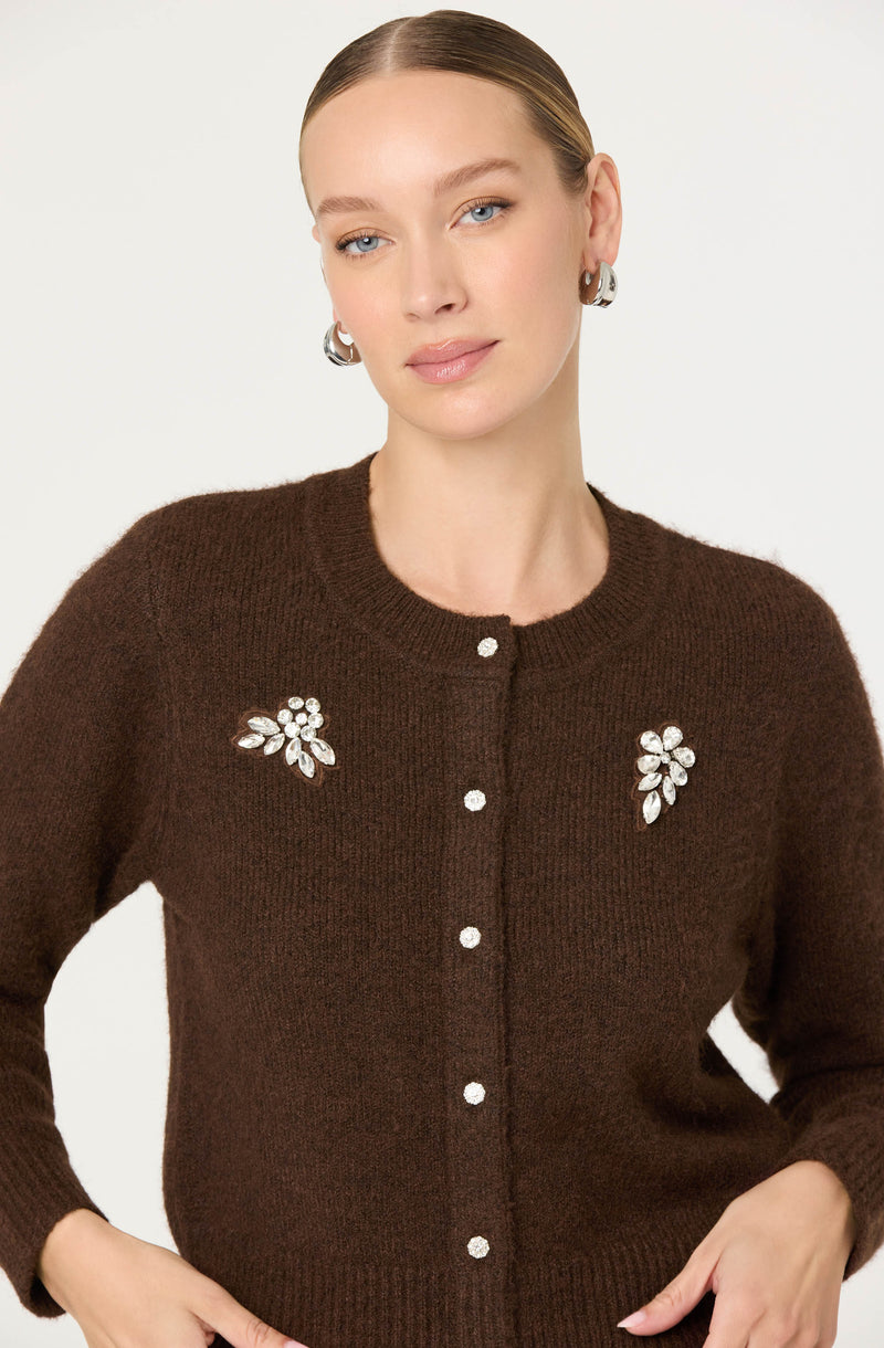 A woman is wearing a dark brown, long-sleeved, button-up cardigan with a round neckline. The cardigan features decorative embellishments of silver-tone floral brooches with rhinestones, positioned on each side of the chest. She has her hair slicked back and is accessorized with large silver hoop earrings.