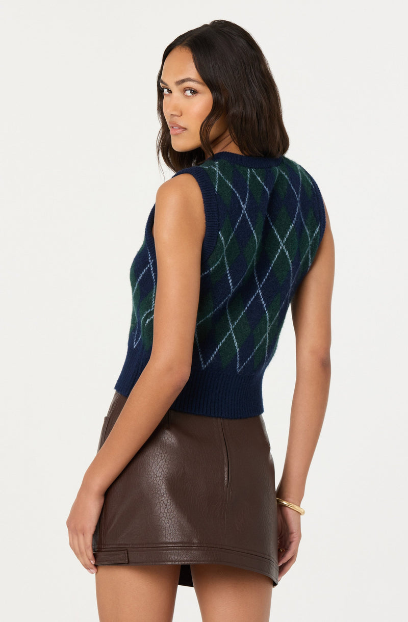 A woman is wearing a sleeveless, navy blue knit sweater vest with an argyle pattern in green, light blue, and white. She is also wearing a brown, leather-looking mini skirt.