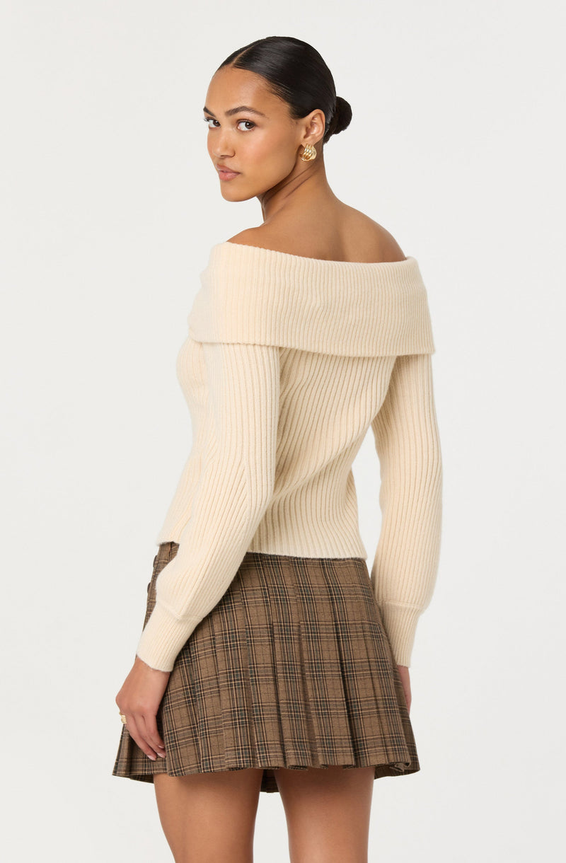 A woman is wearing an off-the-shoulder cream-colored ribbed sweater with a wide, folded neckline that drapes over her shoulders. The sweater has long sleeves and a slim fit. She pairs it with a brown plaid skirt featuring a classic check pattern.