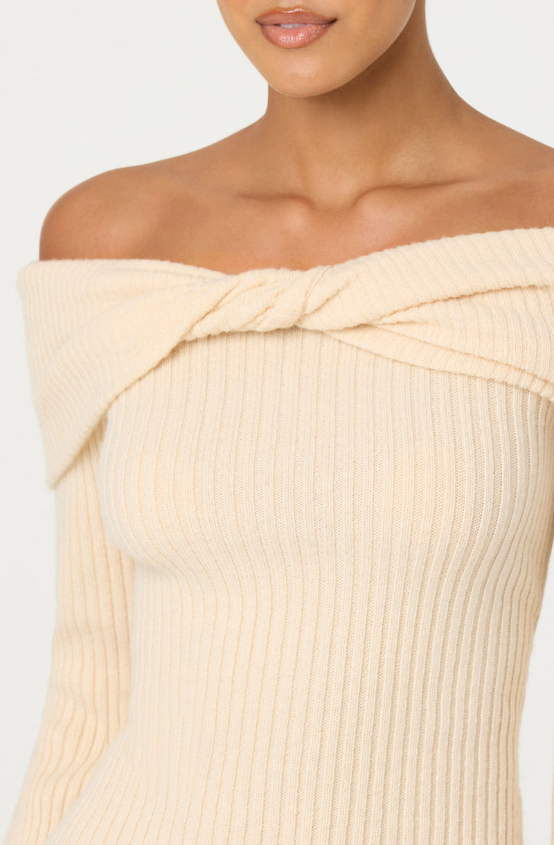 A woman is wearing an off-the-shoulder sweater in a light cream color. The sweater features a ribbed knit texture with vertical lines and a twisted knot detail at the neckline.