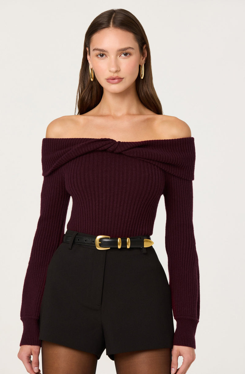 A woman wearing an off-the-shoulder, long-sleeved, ribbed knit sweater in a deep burgundy color. She has on high-waisted black shorts with a black belt featuring a gold buckle. She accessorizes with large gold hoop earrings.