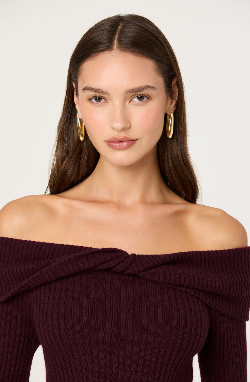A woman with long brown hair is wearing a dark burgundy off-the-shoulder ribbed knit top with a twisted fabric detail at the neckline. She has large gold hoop earrings and a subtle, natural makeup look.