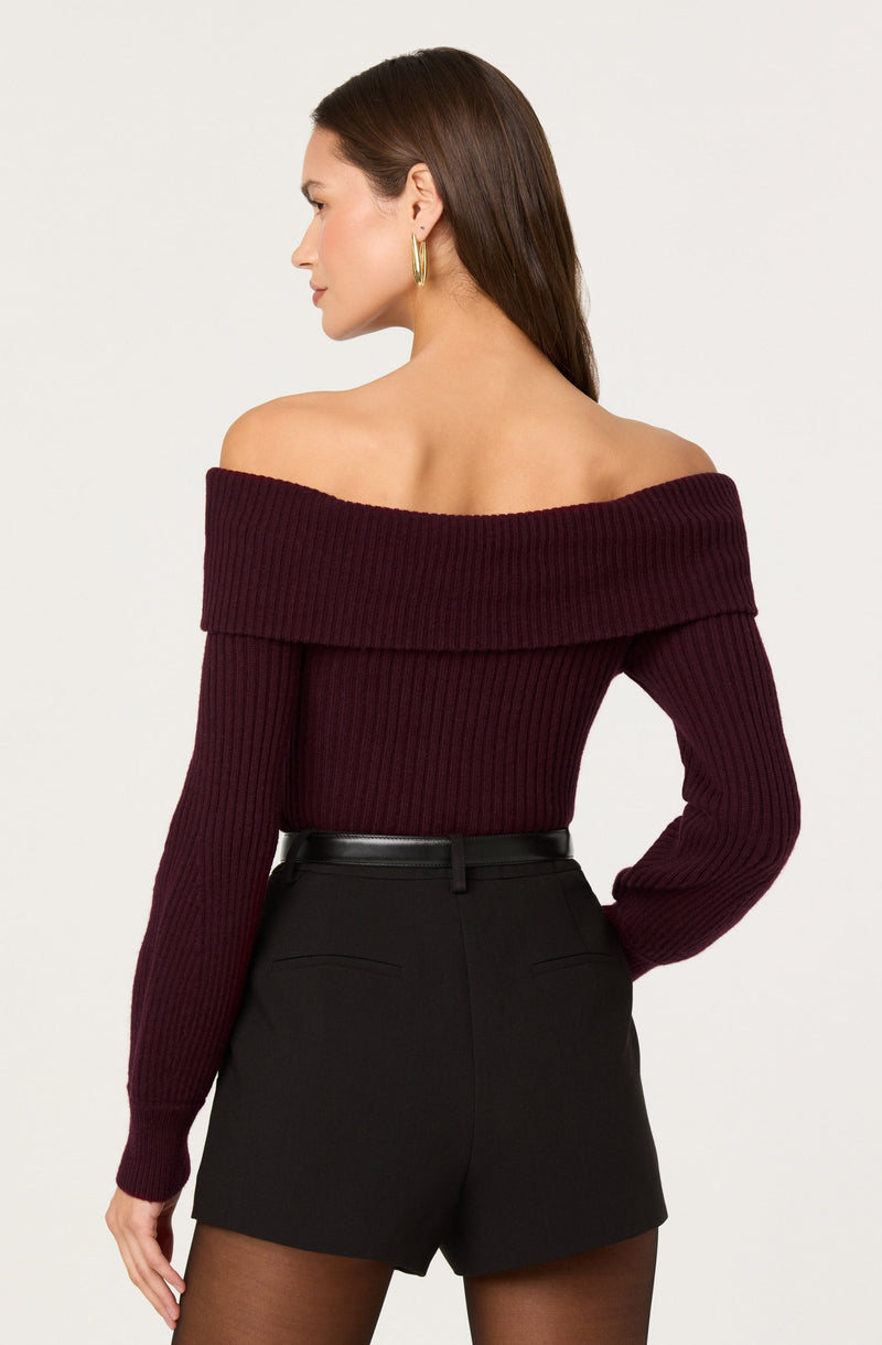 A woman is wearing an off-the-shoulder, long-sleeved, ribbed sweater in a deep burgundy color. She has paired it with high-waisted black shorts and sheer black tights. She accessorizes with large gold hoop earrings and has long, straight brown hair.