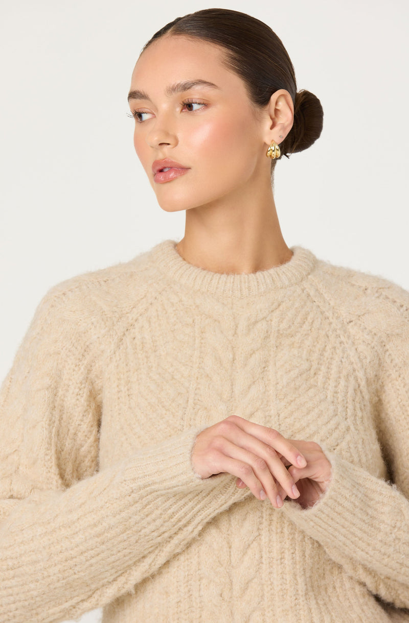 A woman is wearing a cream-colored, chunky knit sweater with a cable knit pattern on the front and ribbed cuffs and neckline. She has gold hoop earrings and her hair is styled in a low bun.