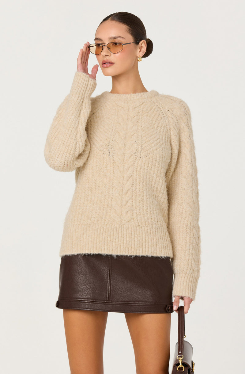 A woman wearing a cream-colored chunky knit sweater with cable knit patterns on the front and a ribbed hem, paired with a dark brown leather mini skirt. She has hair pulled back, wears tinted sunglasses, and has gold earrings. Her right hand is raised near her face, and she holds a small handbag with a strap in her left hand.