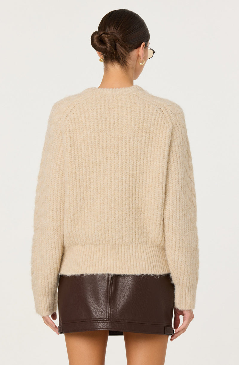 A woman is wearing a cream-colored, chunky knit sweater with a ribbed hem and cuffs. She pairs it with a dark brown, leather mini skirt that has a slight sheen and a small slit at the hem.