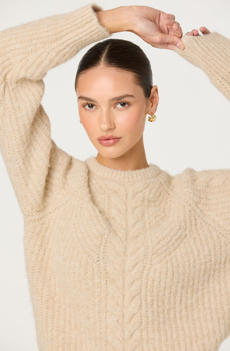 A woman wearing a cozy, cream-colored, chunky knit sweater with a cable-knit pattern down the center. She has gold hoop earrings and her dark hair is pulled back. Her arms are raised above her head, with one hand touching her wrist.