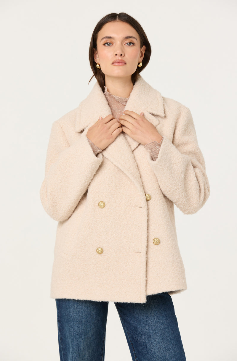 A woman wearing a light beige, double-breasted textured coat with gold buttons. She has dark brown hair, styled straight, and is accessorized with gold earrings. Underneath the coat, she is wearing a light pink or mauve sweater. She is holding the lapels of her coat with both hands and is standing against a plain white background.