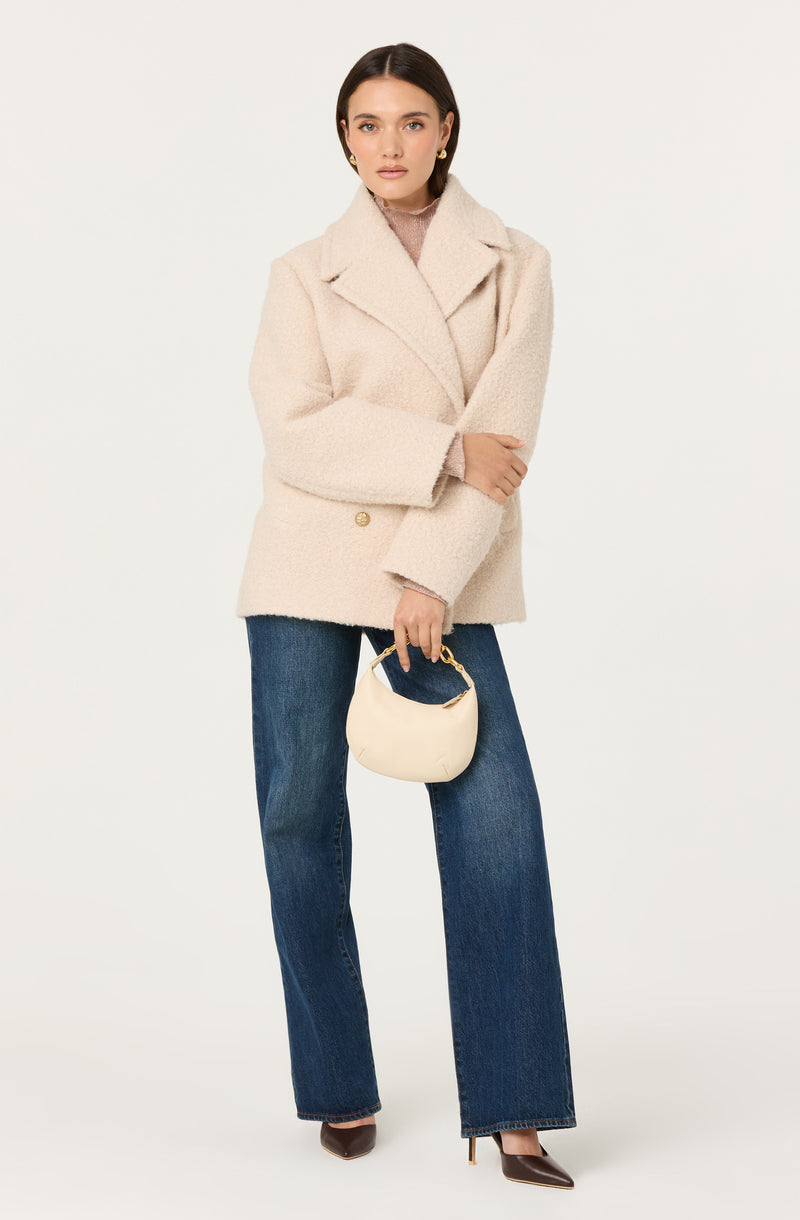 The woman is wearing a light beige, textured, double-breasted coat with wide lapels and long sleeves. She is also dressed in dark blue wide-leg jeans and brown pointed-toe high heels. She is holding a small, cream-colored handbag with a gold clasp.